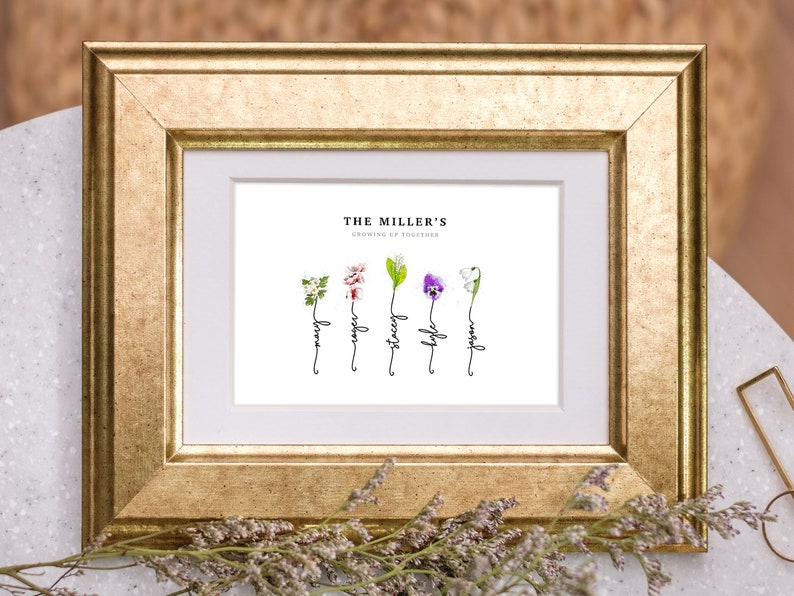 Grandmas Garden personalized birth flower print in a gold frame with white mat, sitting on a marble table with dried flowers. Five watercolor birth month flowers (hawthorn, cherry blossom, lily of the valley, violet and snowdrop) grow from hand-lettered stems that spell each name. Custom family name art and sentimental grandma gift.