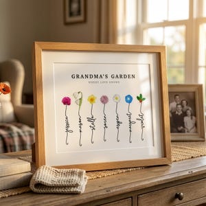 Grandma's Garden personalized birth flower print in a light oak frame with white mat, propped on a dresser in a sunlit living room beside a vintage family photo. Subtitle reads Where Love Grows. Seven watercolor birth month flowers, including carnation, snowdrop, daffodil, allium, daisy, morning glory and holly, grow from hand-lettered stems spelling each grandchild's name. A sentimental grandma gift from the grandkids.