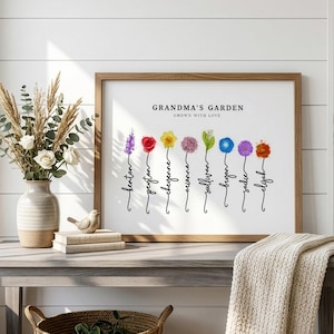 Grandma's Garden personalized birth flower print in a wood frame, leaning on a console table beside white roses, dried grasses and a knit throw. Subtitle reads Grown With Love. Eight watercolor birth month flowers, including larkspur, rose, daffodil, allium, lily of the valley, morning glory, aster and marigold, grow from hand-lettered stems that spell each grandchild's name. A sentimental grandma gift from the grandkids.