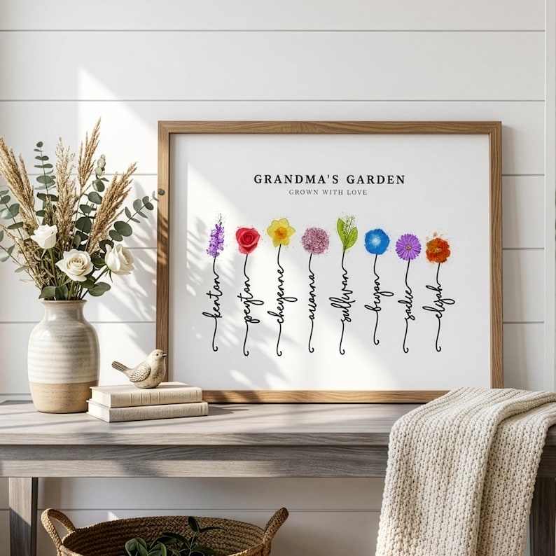 Grandma's Garden personalized birth flower print in a wood frame, leaning on a console table beside white roses, dried grasses and a knit throw. Subtitle reads Grown With Love. Eight watercolor birth month flowers, including larkspur, rose, daffodil, allium, lily of the valley, morning glory, aster and marigold, grow from hand-lettered stems that spell each grandchild's name. A sentimental grandma gift from the grandkids.
