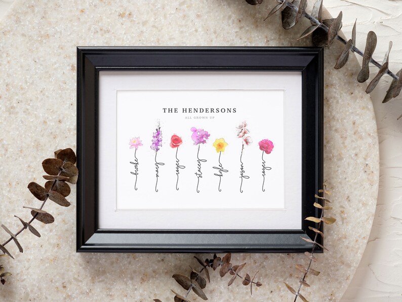 Grandmas Garden personalized birth flower print in a black frame with white mat, styled on stone with dried eucalyptus. Custom family name art titled The Hendersons, All Grown Up, with seven watercolor birth month flowers - water lily, larkspur, rose, sweet pea, daffodil, cherry blossom and peony - each rising from a hand-lettered stem that spells a family member's name. Sentimental grandma gift and birth flower bouquet wall art.