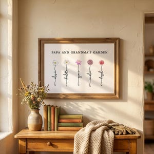Papa and Grandma's Garden personalized birth flower print in a rustic wood frame, hung in a sunlit entryway above a table with dried flowers, books and garden gloves. Five birth month flowers, including narcissus, water lily and allium, grow from hand-lettered stems that spell each grandchild's name. A grandparent gift from the grandkids that names both grandma and grandpa on one print.