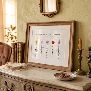 Grandma Ali's Garden personalized birth flower print in a rustic wood frame with white mat, leaning on a marble dresser with lace doilies, brass candlestick and dried rosebuds. Subtitle reads We All Love You So Much. Five watercolor birth month flowers, including daffodil, aster, cosmos, poppy and larkspur, grow from hand-lettered stems spelling each grandchild's name. A personalized grandma gift with her own name on it.