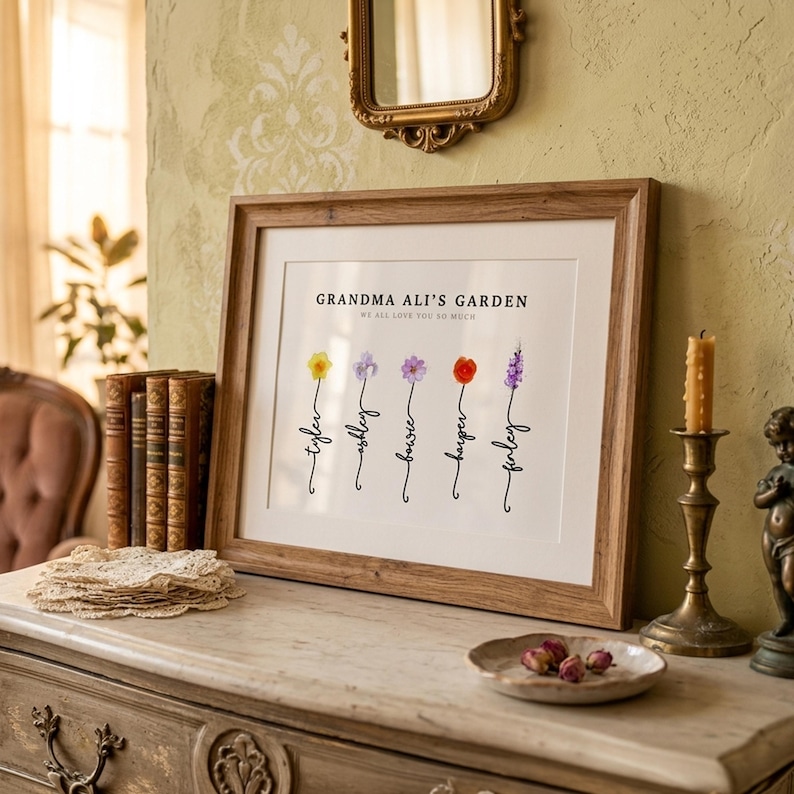 Grandma Ali's Garden personalized birth flower print in a rustic wood frame with white mat, leaning on a marble dresser with lace doilies, brass candlestick and dried rosebuds. Subtitle reads We All Love You So Much. Five watercolor birth month flowers, including daffodil, aster, cosmos, poppy and larkspur, grow from hand-lettered stems spelling each grandchild's name. A personalized grandma gift with her own name on it.