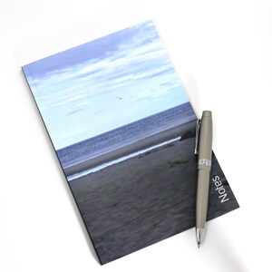 May include: A grey notebook with a photo of a beach and ocean on the cover. A grey pen with the word "Notes" printed on it is lying on the notebook.