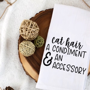 May include: White kitchen towel with black text that reads "cat hair a condiment & an accessory". The towel is on a wooden round slice with brown and beige twine balls.