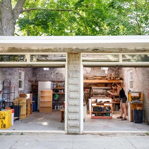 May include: A two-car garage with stone block walls and open bays. The left bay contains a yellow storage bin, a black crate, and a wooden shelf. The right bay features a workbench with various tools, wooden boards, and a black trash can.