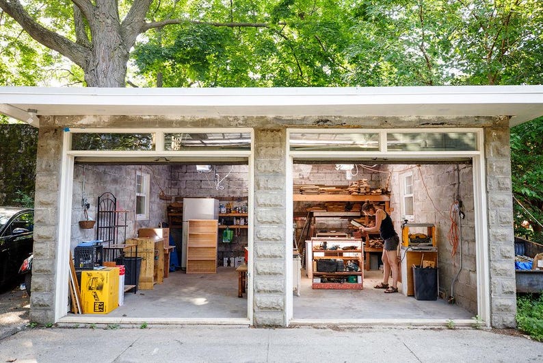 May include: A two-car garage with stone block walls and open bays. The left bay contains a yellow storage bin, a black crate, and a wooden shelf. The right bay features a workbench with various tools, wooden boards, and a black trash can.