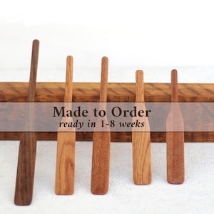 May include: Five wooden spatulas of varying lengths and wood tones are arranged in a row in front of a rectangular wooden block. The spatulas feature long, slender handles and flat, rectangular blades with rounded bottom edges, showcasing natural grain.