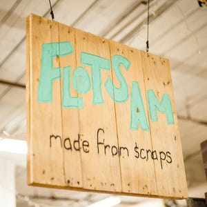 May include: A rectangular wooden sign hanging from thin black wires. The sign features the word FLOTSAM painted in large, light teal letters, with the phrase made from scraps written below in smaller, black, hand-drawn lettering.