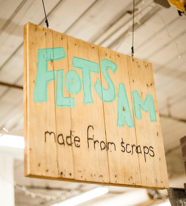 May include: A rectangular wooden sign hanging from thin black wires. The sign features the word FLOTSAM painted in large, light teal letters, with the phrase made from scraps written below in smaller, black, hand-drawn lettering.