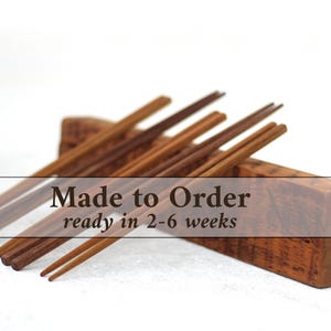 May include: A set of wooden chopsticks resting on a wooden block. The chopsticks are a warm brown color, and the block has a textured, natural wood grain. The image includes the text "Made to Order".