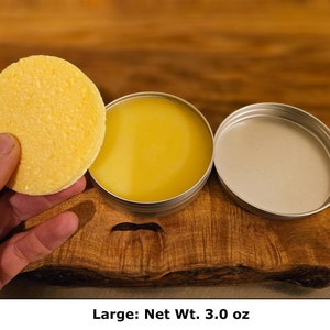 May include: A round, textured yellow solid disc held next to an open circular metal tin containing a smooth, yellow balm. The tin has a separate metal lid resting beside it. The product is labeled as Large with a Net Wt. of 3.0 oz.