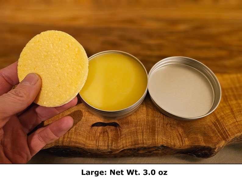 May include: A round, textured yellow solid disc held next to an open circular metal tin containing a smooth, yellow balm. The tin has a separate metal lid resting beside it. The product is labeled as Large with a Net Wt. of 3.0 oz.