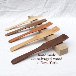 May include: A collection of six wooden spatulas in varying shades of brown, from light tan to dark walnut. The spatulas are handmade from salvaged wood and are arranged on a white surface. Text on the image reads: handmade with salvaged wood in New York.
