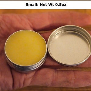 May include: A small round silver metal tin containing a yellow balm, with a matching screw-top lid placed beside it. The text Small: Net Wt 0.5oz is printed above the container, indicating the weight of the product inside the circular tin.