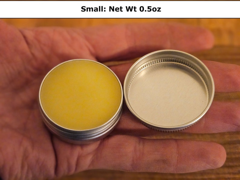 May include: A small round silver metal tin containing a yellow balm, with a matching screw-top lid placed beside it. The text Small: Net Wt 0.5oz is printed above the container, indicating the weight of the product inside the circular tin.