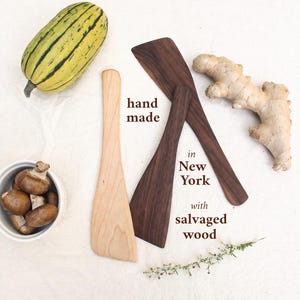 May include: Three wooden spatulas, one light-colored and two dark-colored, arranged with a squash, ginger root, and a bowl of mushrooms. Text on the image reads: hand made in New York with salvaged wood.