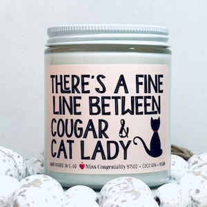 May include: A white candle jar with a pink label that reads "THERE'S A FINE LINE BETWEEN COUGAR & CAT LADY". The label also includes a black silhouette of a cat with a pink collar.