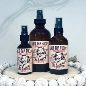 May include: Three glass spray bottles with black tops and labels that read "Shut Tha Fucup & Smell This". The labels feature a cartoon woman with a finger to her lips. The bottles are on a wooden surface with white beads.