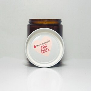 May include: A brown glass jar with a white lid. The lid has a pink heart-shaped sticker with the text "Miss Congeniality SCENT CHOICE".