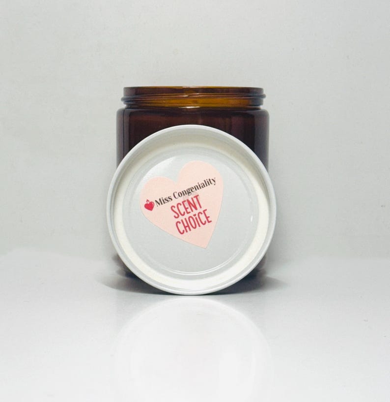 May include: A brown glass jar with a white lid. The lid has a pink heart-shaped sticker with the text "Miss Congeniality SCENT CHOICE".
