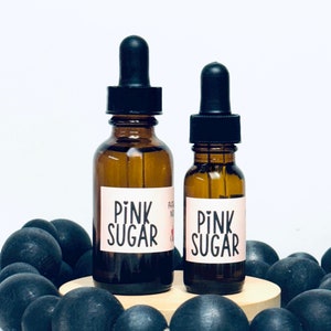 May include: Two amber glass bottles with black dropper tops and labels that say "Pink Sugar". The bottles are sitting on a wooden platform surrounded by black beads.