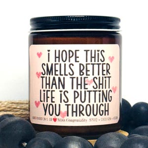 May include: A brown glass candle jar with a white label that reads "I hope this smells better than the shit life is putting you through". The label also includes pink hearts and the text "Hand poured in S. OR Miss Congeniality 97502 COCO SOY WEGUN".