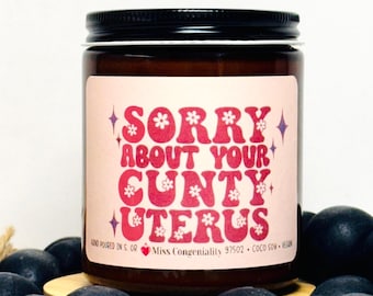 May include: A brown glass candle jar with a pink label that reads "SORRY ABOUT YOUR CUNTY UTERUS". The label has a pink and white floral design. The candle is hand-poured in the USA and made with coconut soy wax.