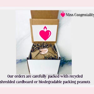 May include: A white box with a brown lid is filled with shredded paper. The lid has a pink heart with a flame on top and the text "Miss Congeniality". The text "Our orders are carefully packed with recycled shredded cardboard or biodegradable packing peanuts" is below the box.
