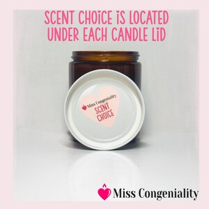 May include: A brown glass candle jar with a white lid. The lid has a pink heart with the text "Miss Congeniality Scent Choice" on it. The text "Scent Choice is located under each candle lid" is written in black text on a pink background.