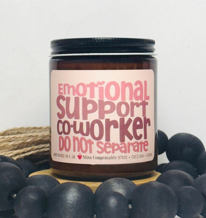 May include: A brown glass candle jar with a white label that reads "Emotional Support Co-worker DO NOT separate". The label also includes the text "Hand poured in S. OR Miss Congeniality 97502 COCO SOY VEGAN".