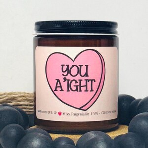 May include: A brown glass candle jar with a pink heart-shaped candy design and the text "you a'ight". The jar has a black lid and is sitting on a wooden surface.