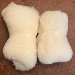 May include: Two bundles of off-white, fluffy raw wool fiber. Each bundle is secured with a thin, light-colored string tied around the center, creating a soft, cloud-like appearance with a fibrous, textured surface throughout the material.
