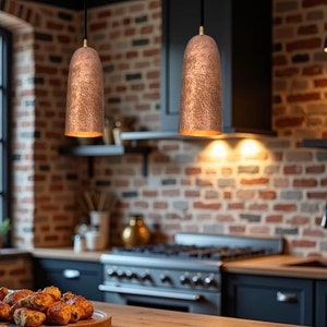 May include: Two copper pendant lights hang over a kitchen counter with a brick wall backdrop. The lights are on and casting a warm glow. A wooden cutting board with baked goods sits on the counter.
