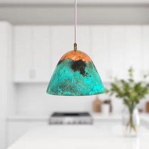 Puede incluir: Una l&aacute;mpara colgante turquesa y cobre con un acabado &uacute;nico y desgastado. La luz cuelga de un cable blanco en un entorno de cocina.