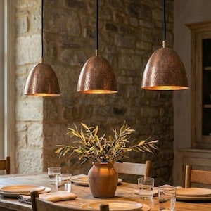 Peut inclure: Trois lampes suspendues en cuivre avec une texture martelée sont suspendues au-dessus d'une table à manger en bois. Les lampes sont suspendues par des cordons noirs et dégagent une lueur chaleureuse. Un vase de verdure est posé sur la table.