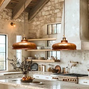Puede incluir: Dos lámparas colgantes de cobre cuelgan sobre una isla de cocina. Las lámparas tienen una textura martillada y un acabado metálico cálido. La cocina presenta paredes de piedra, estantes de madera y una encimera de mármol, creando un ambiente rústico y acogedor.