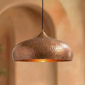 Op de afbeelding: Een koperkleurige hanglamp met een gehamerde textuur. De lamp heeft een koepelvorm en een warme, uitnodigende gloed. De lamp hangt aan een zwart snoer.