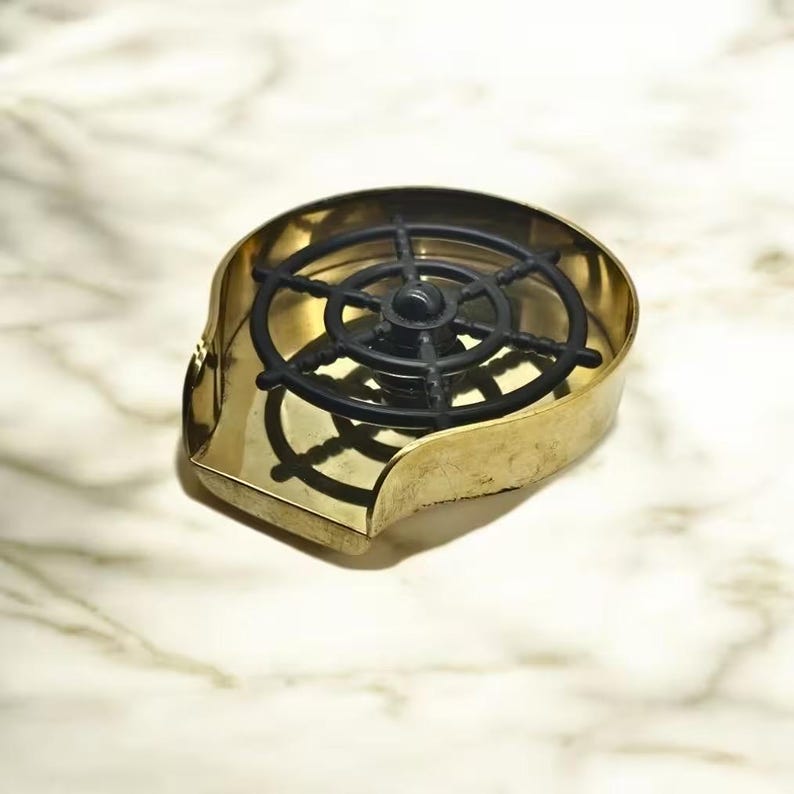 May include: A gold-colored soap dish with a black plastic wheel-shaped soap holder. The dish is shaped like a seashell.