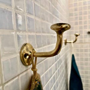 May include: A gold-toned metal hook mounted on a white tiled wall. The hook has a rounded top and a curved arm. A brown fabric bag hangs from the hook.