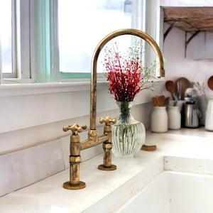 Może przedstawiać: Złota bateria kuchenna z dwoma uchwytami i wysokim wylewką. Bateria jest zamontowana na białym blacie. W tle znajduje się wazon z czerwonymi kwiatami.