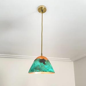 Puede incluir: Una l&aacute;mpara colgante de cobre con un acabado de p&aacute;tina verde y marr&oacute;n. La luz tiene una pantalla en forma de c&uacute;pula y un cable de color dorado.