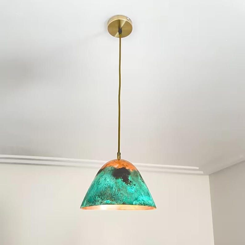 Puede incluir: Una l&aacute;mpara colgante de cobre con un acabado de p&aacute;tina verde y marr&oacute;n. La luz tiene una pantalla en forma de c&uacute;pula y un cable de color dorado.