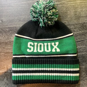 May include: Black knit beanie with a green and white striped band and a green pom pom. The word "SIOUX" is written in white letters on the green band.