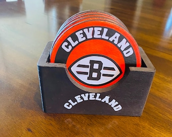 May include: A set of six round coasters with a black and white Cleveland Browns logo on a red background. The coasters are in a black wooden holder with the words "Cleveland" on the front.