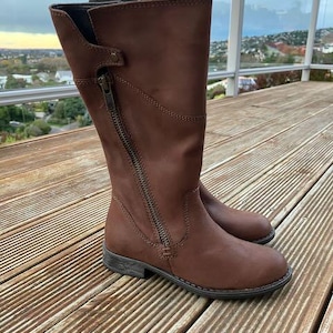 May include: A pair of brown leather riding boots with a side zipper. The boots have a low heel and are knee-high. The boots are sitting on a wooden deck with a view of a cityscape in the background.
