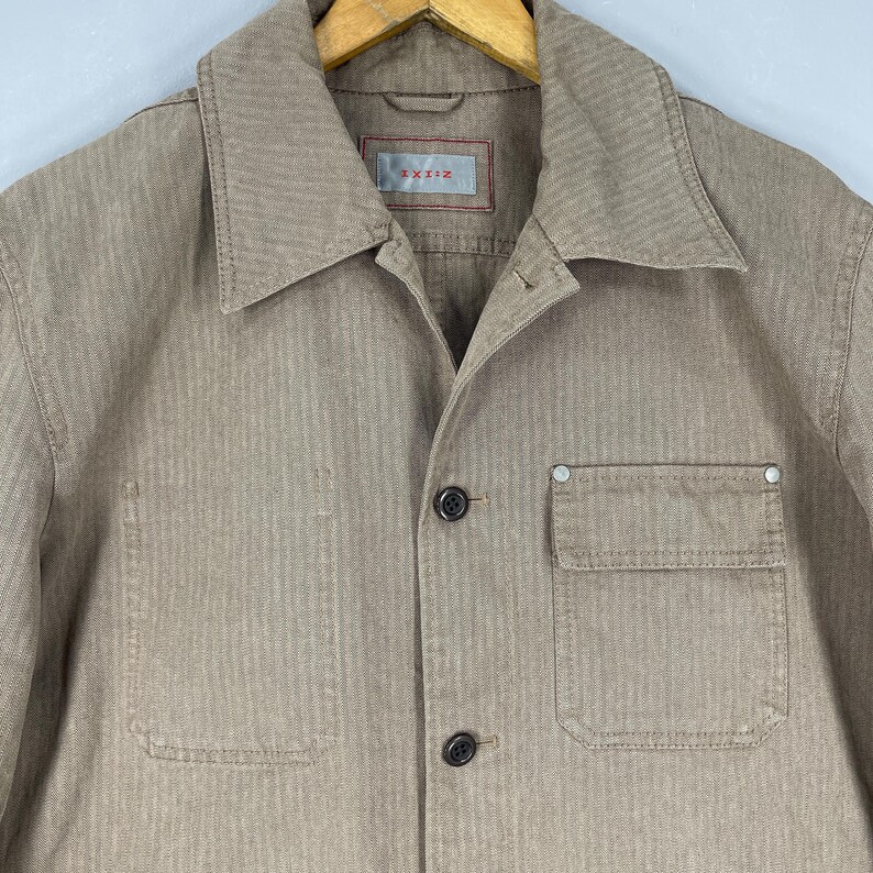 May include: Tan button-up shirt with a collared neckline and a single chest pocket. The shirt has a textured, woven appearance and features the text "TXI:Z" on a label near the collar.