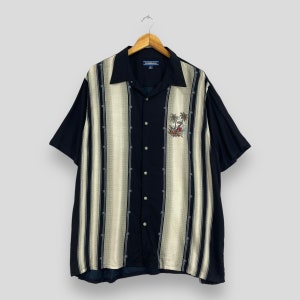 Puede incluir: Una camisa de manga corta con botones, a rayas blancas y negras, con un diseño de palmeras. La camisa tiene cuello y un bolsillo en el lado izquierdo. La marca "Caribeana" es visible en la etiqueta.