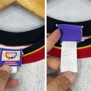Puede incluir: Primer plano de una sudadera gris de cuello redondo con cuello negro y rayas rojas y amarillas. La etiqueta dice "SERIOUS IMPACT LEGENDS" en un rect&aacute;ngulo azul.